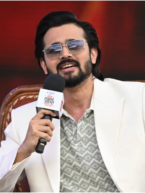 Bhuvan Bam in a white blazer wearing silver square aviator sunglasses, speaking at a public event.